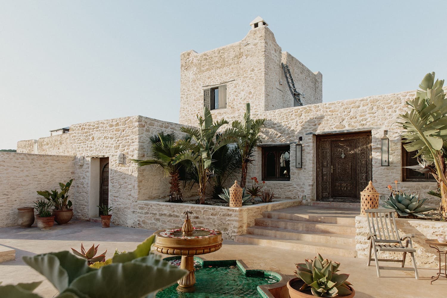 Villa Anouk in Essaouira with a stone facade, ornate door, palms, and a small fountain.