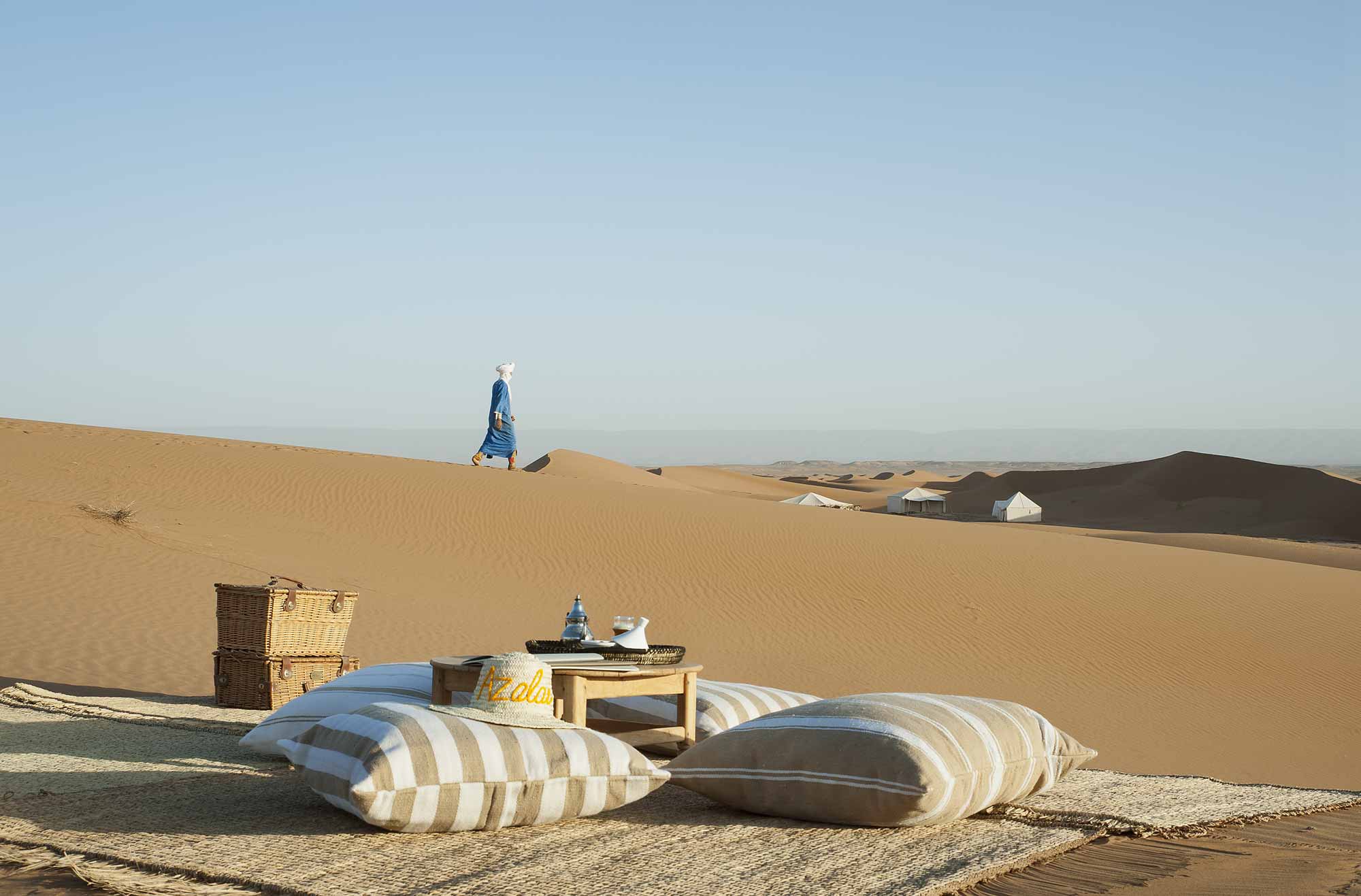 Amidst the rolling sand dunes, an elegant outdoor relaxation spot is arranged with striped cushions and a tea service, while a person in traditional dress strolls by the Azalai Desert Camp.