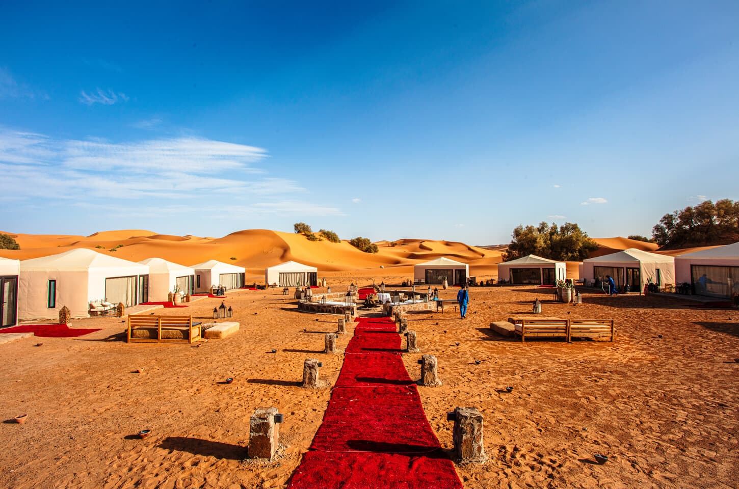 The Merzouga Luxury Desert Camp is set amidst vast sandy terrain, featuring elegant white guest tents, outdoor seating areas, and a welcoming red carpet leading through the camp.