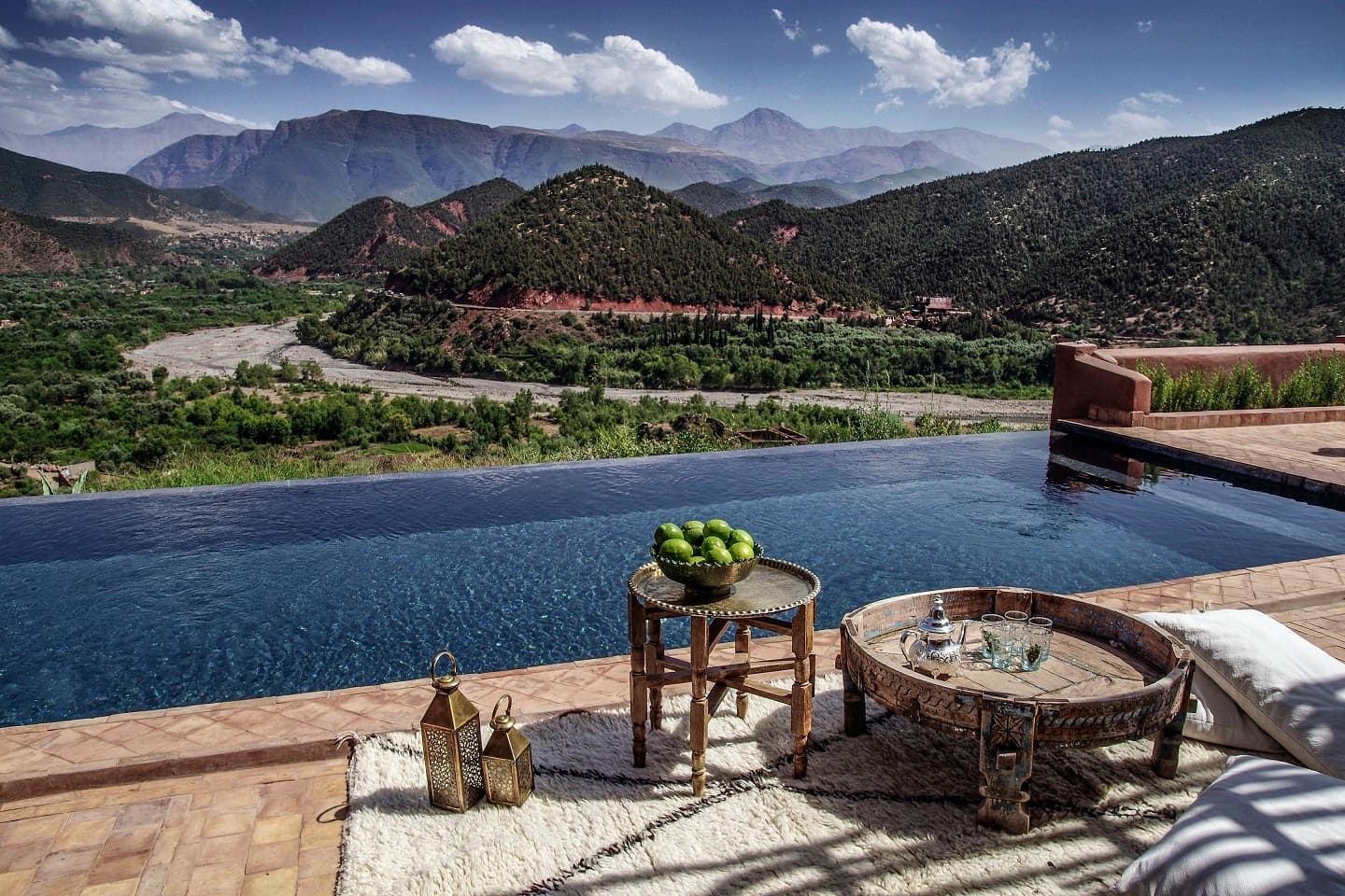 An infinity pool at Kasbah Bab Ourika offers a breathtaking view of the Atlas Mountains, accompanied by a traditional Moroccan side table with a silver teapot set and a bowl of fresh limes.