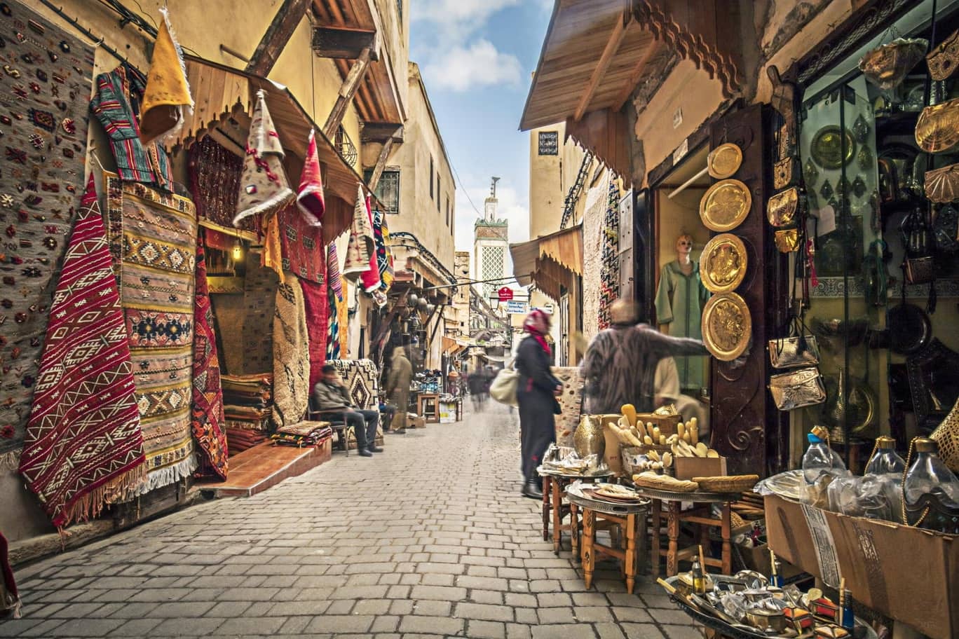 a cooking class on a charming rooftop in Fes, Morocco. Participants, some wearing traditional Moroccan caps, are preparing ingredients at their individual stations surrounded by lush potted plants and vibrant red walls in the background.