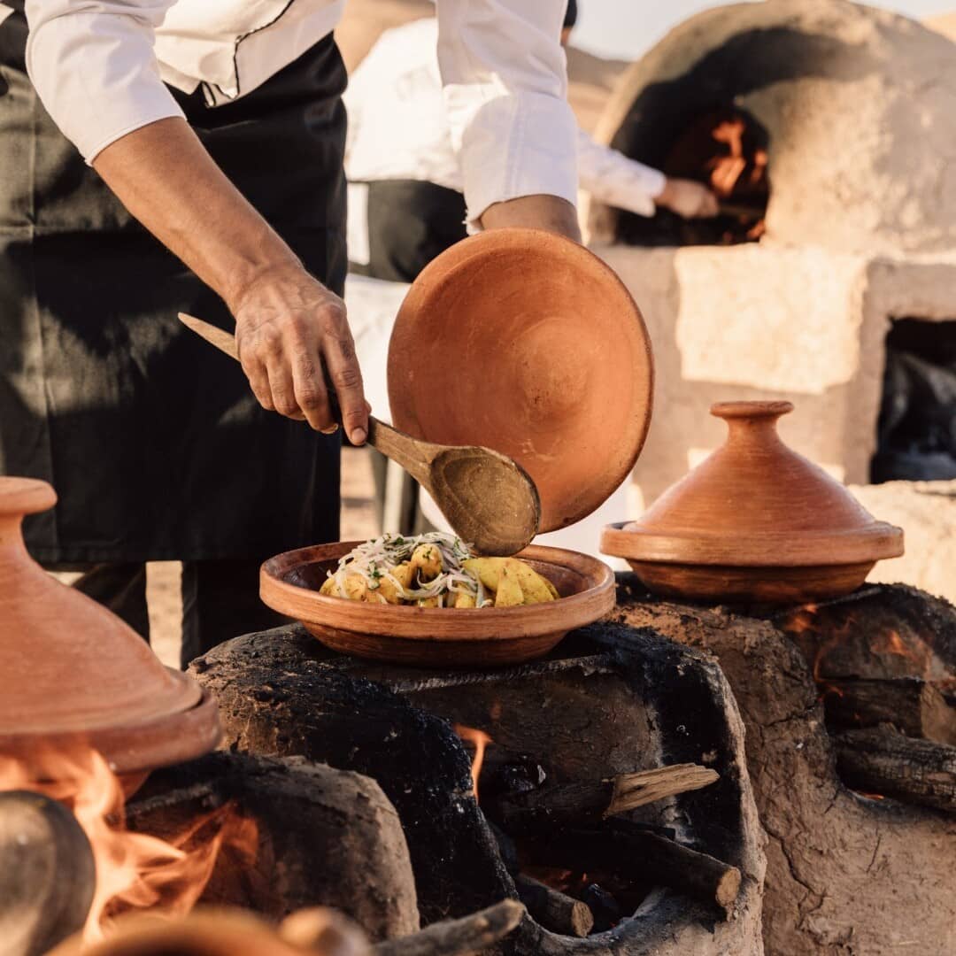 A chef artfully prepares a traditional Moroccan dish in a tagine, using an open fire to infuse the ingredients with rich, smoky flavors.