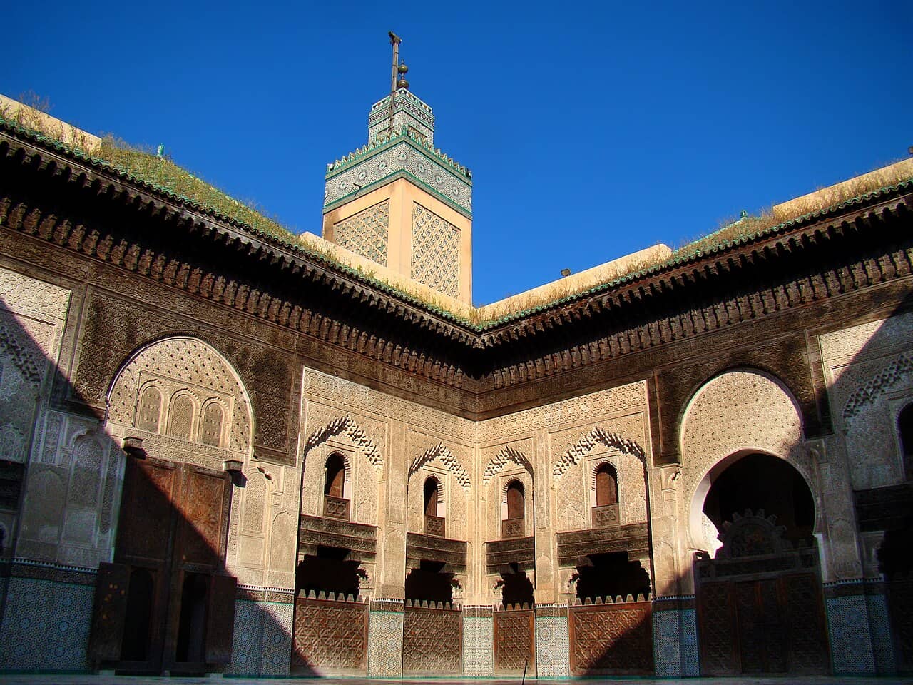  the intricate facade of The Bou Inania Madrasa in Fes, Morocco. The architectural detail includes elaborate carvings and tilework under a deep blue sky. The minaret of the madrasa stands tall in the background, adorned with ornate geometric patterns.