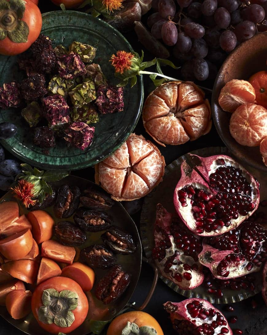 Cover of a travel guide about Moroccan cuisine featuring a close-up of a colorful assortment of Moroccan fruits and sweets, including pomegranates, dates, persimmons, tangerines, and nougat, arranged artfully on a dark background.