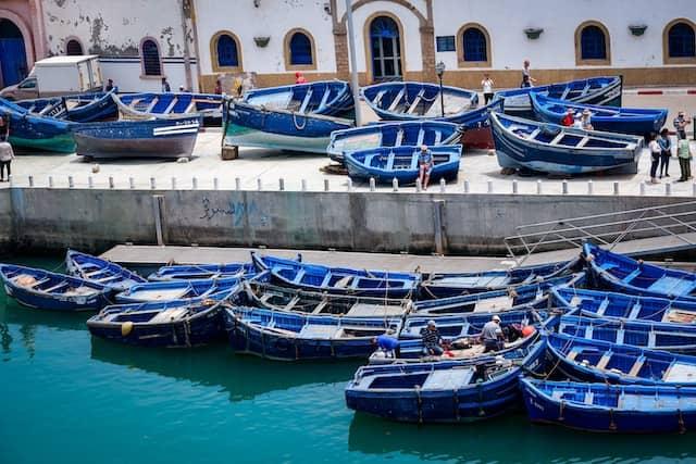 Blue fishing boats docked at a port, Morocco, with white buildings in the background.