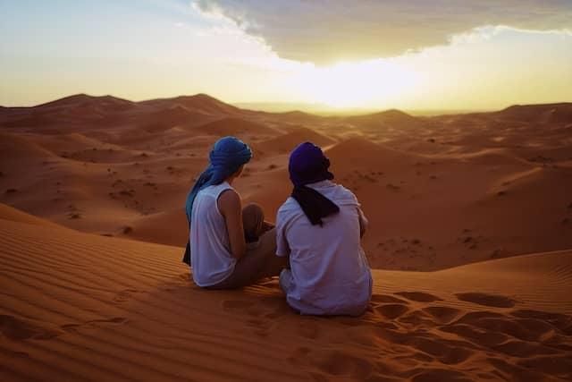 Two people wearing headscarves sitting on a sand dune, watching the sunset in the Sahara Desert.