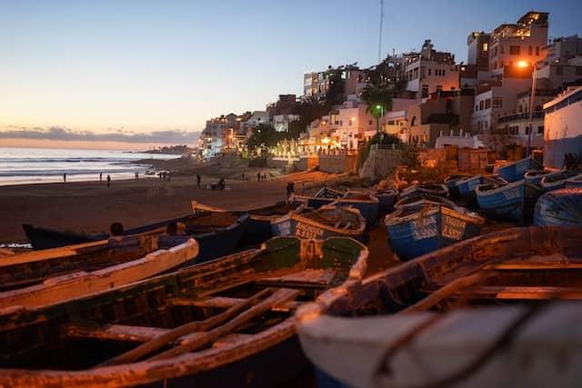 Wooden fishing boats on the shore at sunset with the coastal town of Taghazout, Morocco in the background.