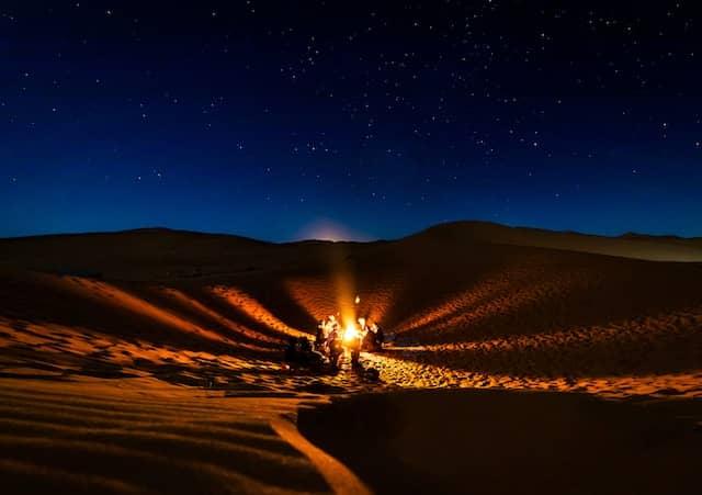 A group of people gathered around a campfire in the Sahara Desert under a starry night sky.