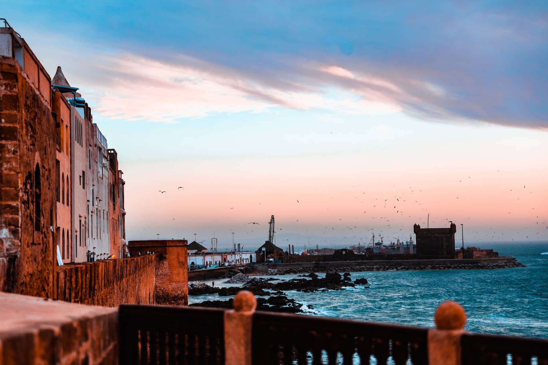 Sunset view of Essaouira's fortified seafront, with historic buildings and flying seagulls against a pastel sky.