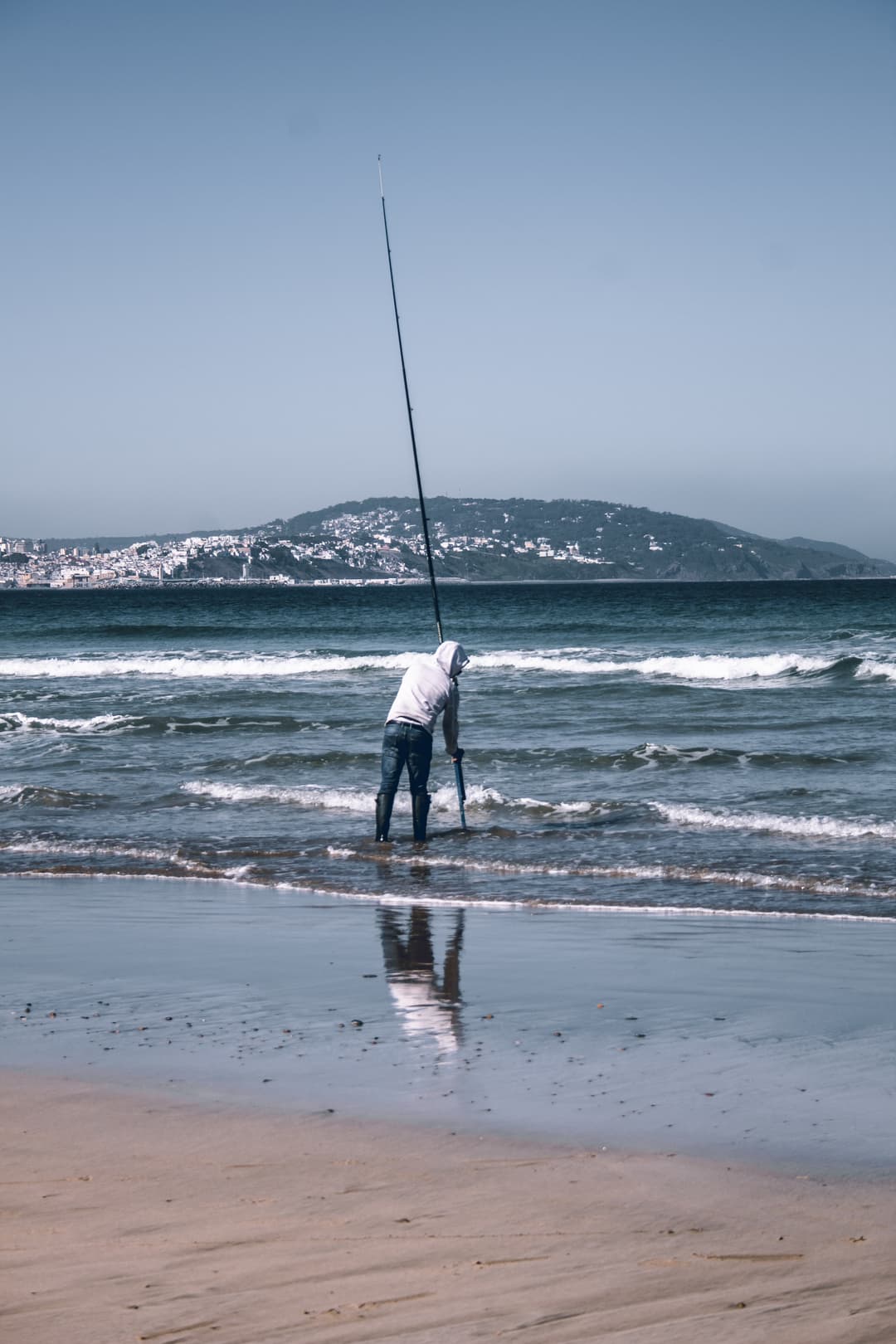 A lone fisherman clad in a hoodie and jeans stands in the shallows on a Tangier beach, fishing rod in hand, with the city's hillside buildings in the distance.